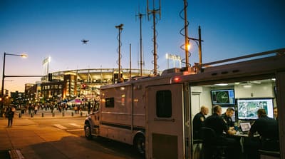 Public safety command vehicle with antenna arrays monitoring low-altitude airspace over a stadium district at dusk, small drone visible in the distance